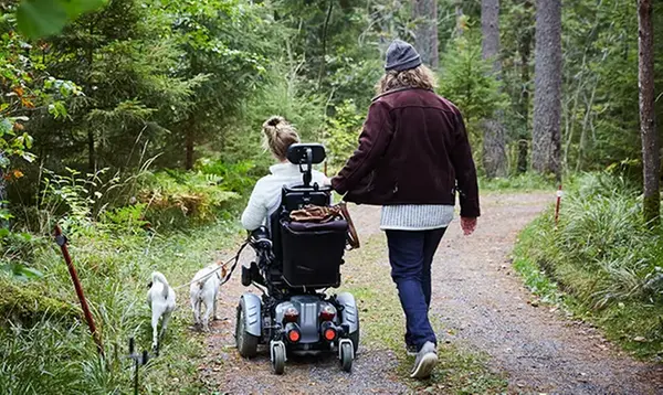 En person i rullstol och en gående person promenerar i skogen